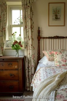 a bed sitting next to a wooden dresser under a window with flowers on the curtains