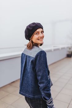 a young woman wearing a denim jacket and beanie smiles while standing on the sidewalk