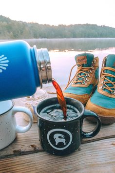 a pair of hiking boots is being poured into a coffee mug on a dock by the water