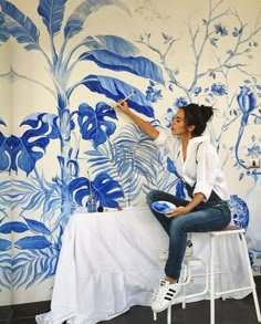 a woman sitting on top of a stool painting a wall with blue flowers and leaves