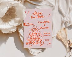 an orange and pink save the date card next to some flowers on a white surface