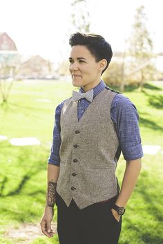 a woman wearing a vest and tie standing in front of a green field with trees