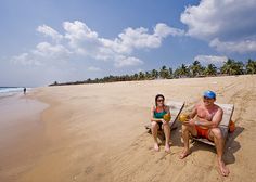 a man and woman sitting on top of a beach next to the ocean with palm trees in the background