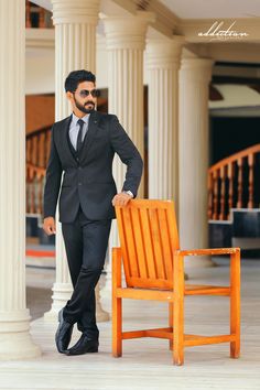 a man in a suit and tie standing next to an orange chair with his hand on the back