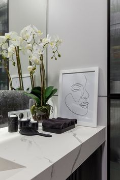 a bathroom counter topped with a vase filled with white flowers next to a mirror and hair dryer