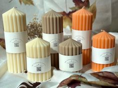 several candles are lined up on a table with autumn leaves in the background and one candle is orange