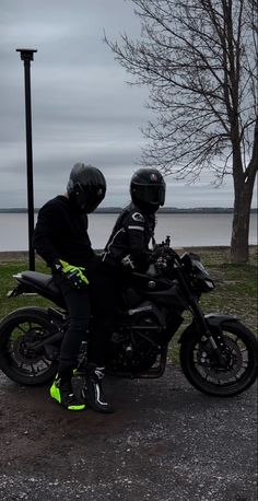 two people sitting on a motorcycle in front of a lake and trees with no leaves