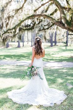 a woman in a wedding dress holding a bouquet and looking down at the ground with trees behind her