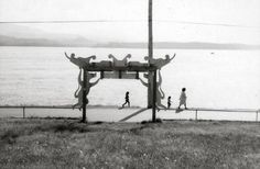 black and white photograph of people walking on the side of a body of water with a sculpture in the foreground