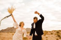 a bride and groom are holding hands in the desert with their arms up as they walk