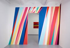 an art gallery with colorful striped walls and white flooring, one room is open to the other