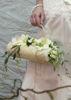 a woman is holding a basket with flowers and greenery on the handle, while wearing a white dress