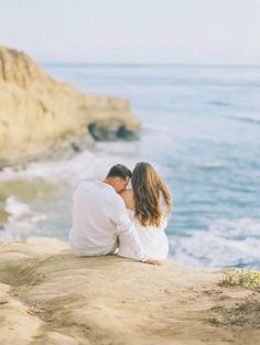 a man and woman sitting on top of a cliff next to the ocean with their arms around each other