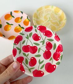 three plates with cherries and oranges painted on them are being held by a hand