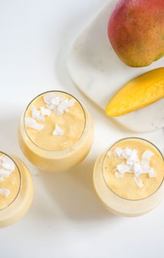 three glasses filled with mango smoothie on top of a white table next to fruit