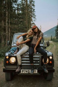 two young women sitting on the back of a black truck in the middle of a forest