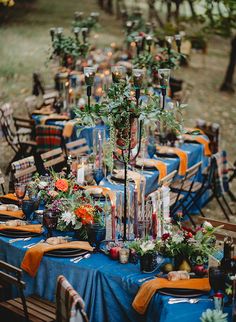 an outdoor dinner table set up with blue linens and orange napkins, candles and greenery