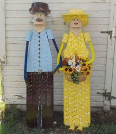 two wooden figures standing next to each other in front of a white building with flowers