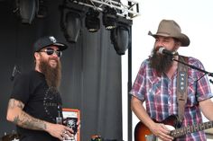 two men with beards are playing guitars and singing into microphones at an outdoor concert