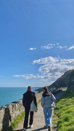 two people walking down a path next to the ocean on a sunny day with blue skies