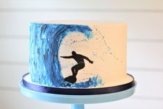 a white and blue cake with a surfer on it's icing is sitting on a stand