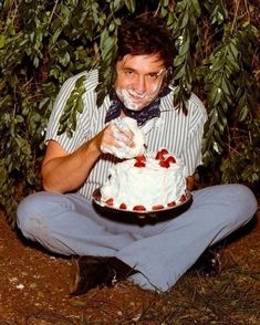 a man sitting on the ground with a cake in front of his face and trees behind him