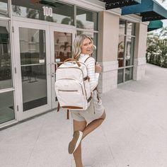 a woman with a white backpack and tan boots is posing for the camera in front of a building