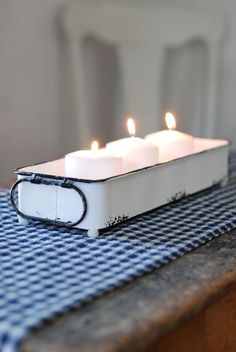 three lit candles in a white container on a blue checkered tablecloth with two chairs behind it