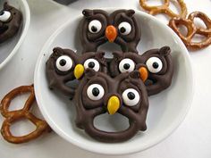 three chocolate owl pretzels with googly eyes on a plate next to pretzel rings