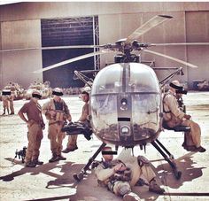 a helicopter that is sitting on the ground with some men around it and another man laying down in front of it