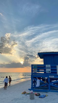 two people are walking on the beach near a blue lifeguard tower at sunset with clouds in the sky