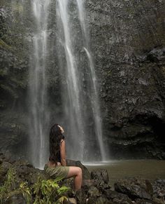 a woman is sitting on rocks in front of a waterfall and looking up at the water