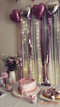 a table topped with a cake covered in pink frosting next to balloons and confetti