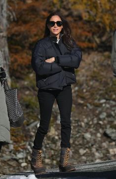 a woman in black jacket and sunglasses standing on snow covered ground with her arms crossed