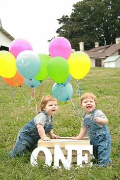 two young boys sitting in front of a wooden one sign with balloons attached to it