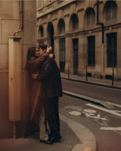 two people standing next to each other on the side of a road with buildings in the background