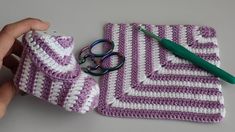 a hand holding a pair of scissors next to a purple and white crocheted bag