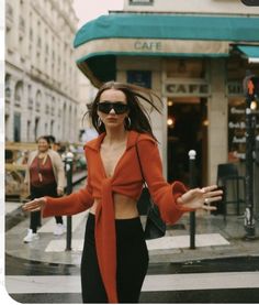 a woman in an orange sweater and black pants is walking down the street with her arms outstretched