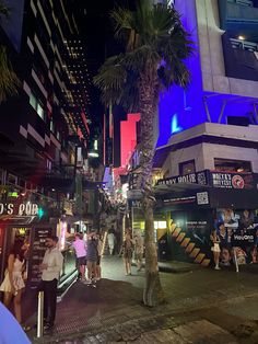 people are standing on the sidewalk in front of some buildings and palm trees at night