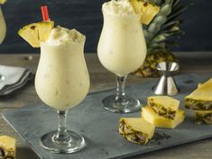 two glasses filled with pineapple milkshakes on top of a slate tray next to sliced pineapples