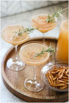 three cocktails with rosemary garnish sit on a wooden tray next to two drinks