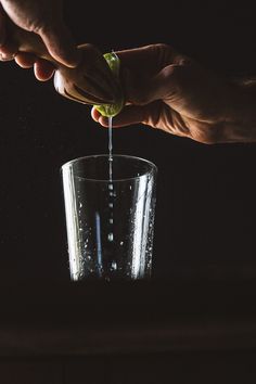 someone is pouring something into a glass with water on the side and another hand reaching for it