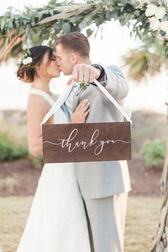 a bride and groom kissing under a wooden thank you sign