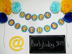 a happy birthday banner and decorations on a white table with blue, yellow and orange pom poms