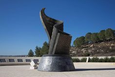 a large metal sculpture sitting on top of a cement slab next to a park filled with trees