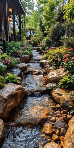 a stream flowing through a lush green forest filled with rocks and flowers next to a house