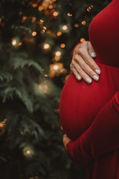 a pregnant woman's hands on her belly in front of a christmas tree with lights