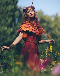 a woman in a red dress with horns and flowers on her head is standing in the middle of a field