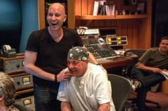 two men laughing in front of a recording studio with other people sitting at the desk