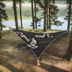 two people laying in a hammock on the ground near some trees and water